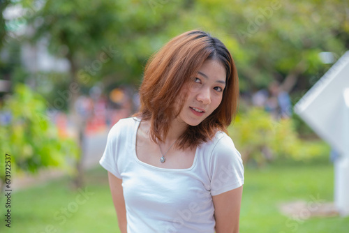 A petite teenage girl with a cute face, white skin, wearing a white t-shirt. Turn to look at the camera and gesture towards the camera. Looks charming and interesting