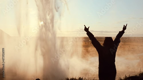 Traveler in silhouette, arms raised, poses for a spectacular sunset photo behind a cascading waterfall. Epic and unforgettable moments. Tourism in natural and spectacular places.