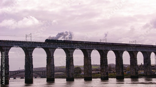 Fototapeta Naklejka Na Ścianę i Meble -  train passing over bridge on river next to town and hills