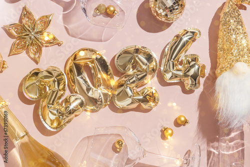 Happy New Year festive background, golden inflatable balls in shape 2024 on pale pink background, sparkling wine champagne bottle, wine glasses with Christmas holiday shiny decor, glitter