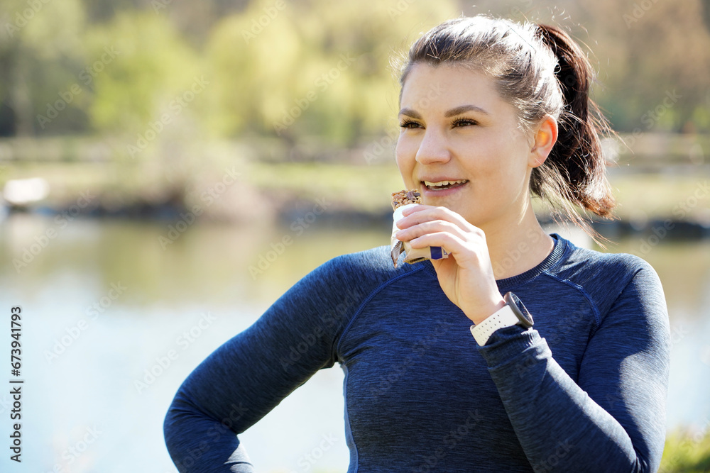 Athletic woman after running and training eats protein bar for muscle building and as an energy booster outdoors in a park with lake on a sunny morning in autumn