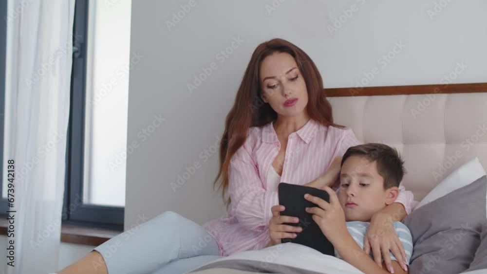 A boy lies on the bed and gets ready to sleep, reading an ebook on a tablet. A young and caring mother sits next to son and strokes his head with careness. The concept of responsiveness in the family. A boy lies on the bed and gets ready to sleep, reading an ebook on a tablet. A young and caring mother sits next to son and strokes his head with careness. The concept of responsiveness in the family.