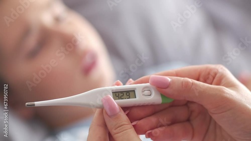 A woman holds a thermometer in her hands. A mother measures and checks son temperature. A sick boy lies on the bed and coughs in the background. Close up.