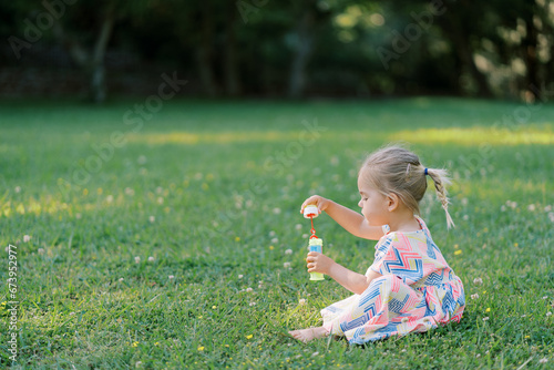 Little girl is sitting on a green meadow with a bottle of soap bubbles in her hands. Side view