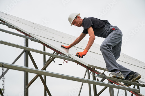 Man technician building photovoltaic solar panels. Male worker in safety helmet fixing solar module on supporting structure while mounting photovoltaic panel system.