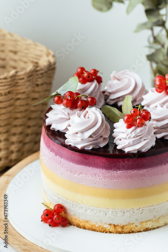 Summer cake with different mousse berry layers decorated with red currant and whipped cream on the wooden cake stand.