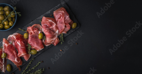 Appetizer from dry cured serrano ham or Spanish jamon iberico. Italian prosciutto crudo served with green olives on the black slate stone board. Meat slices on the black background