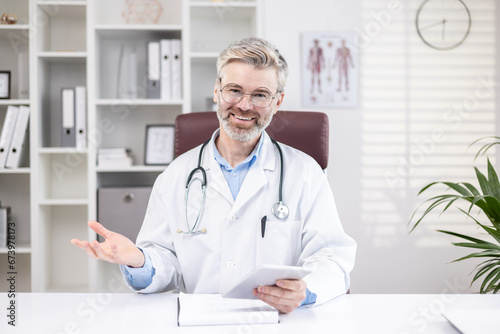 Mature experienced doctor with gray hair, smiling and looking at camera, consulting patients online remotely, senior man working remotely at workplace inside office.