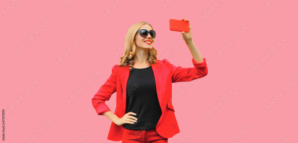Portrait of beautiful elegant lady woman taking selfie with smartphone wearing red business blazer on pink studio background