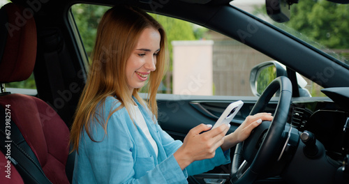 Cheerful blonde girl with freckles sitting in modern car and using mobile phone for surfing internet social media e-mail. Driver checking email chats reading news. Driver writing message SMS vehicle © serg