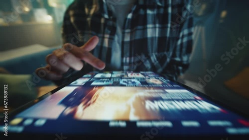 Shot of female hipster wearing checkered shirt holding digital tablet in hands. Young unrecognizable woman choosing TV show to watch on streaming platform. Blurred background of living room. Indoors