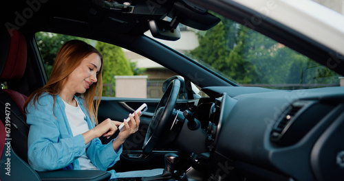Cheerful blonde girl with freckles sitting in modern car and using mobile phone for surfing internet social media e-mail. Woman using touch screen in car setting up the navigation gps application on