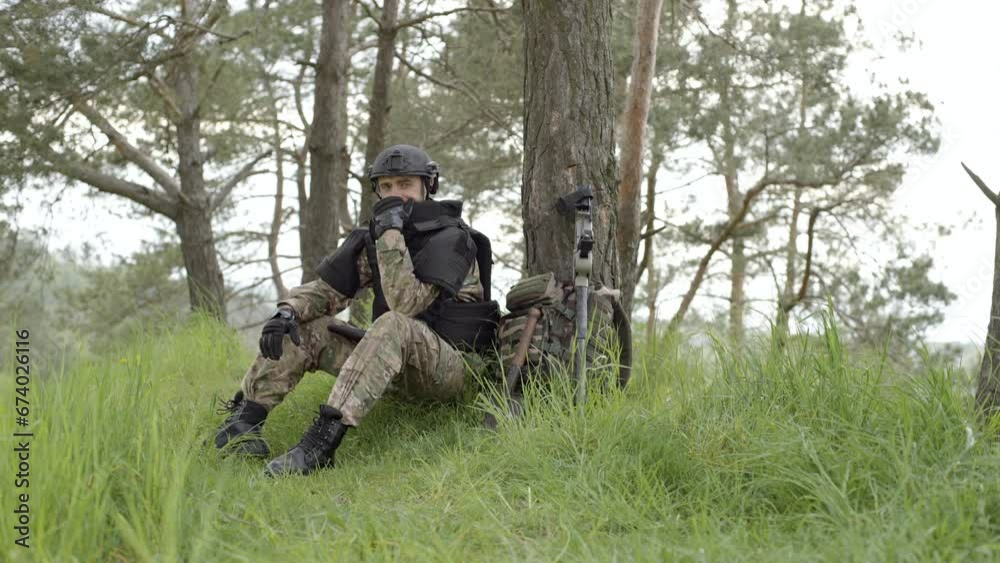 Mine clearance. Soldier with mine detector and backpack sits exhausted by a tree after heavy demining Mine clearance. Soldier with mine detector and backpack sits exhausted by a tree after heavy demining