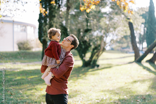 Dad pulls his lips to kiss a little girl in his arms while standing in the park