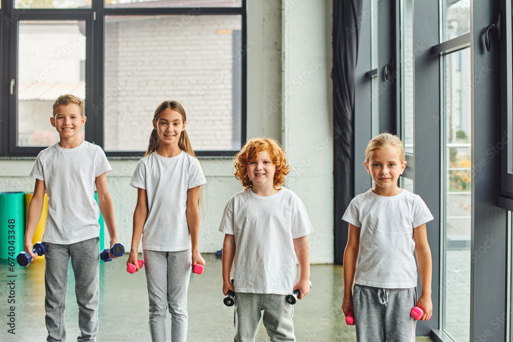 Obraz premium four cheerful little children posing with dumbbells and smiling joyfully at camera, child sport