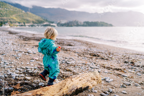 Little girl is walking along a pebbly beach near a driftwood. Side view