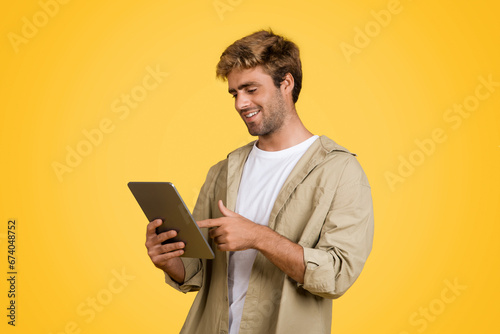 Joyful European man surfing web on tablet, yellow backdrop