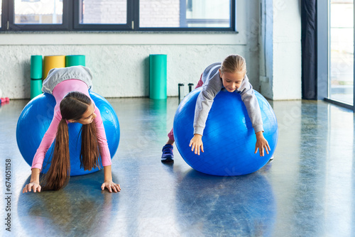 two joyful girls in sportswear lying on fitness balls in gym stretching hands to floor, child sport photo