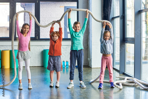 Fototapeta Naklejka Na Ścianę i Meble -  cheerful kids in sportswear raising fitness rope above heads and smiling at camera, child sport