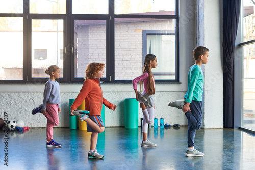 Fotografie four preadolescent pretty children in sportswear stretching their legs in gym, c