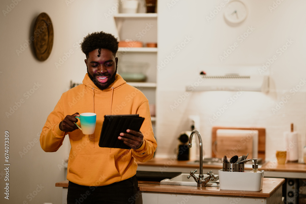 A happy interracial casual millennial is holding a mug of tea and watching the tablet while standing at home at night. A happy interracial casual millennial is holding a mug of tea and watching the tablet while standing at home at night.