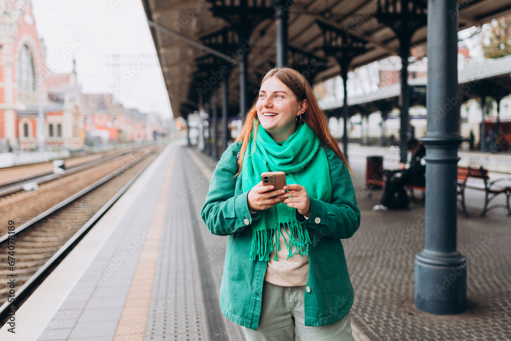 Fototapeta premium A young and beautiful woman with backpack waiting for a train while using a mobile phone. Passenger using cellphone monitoring traffic schedule via application