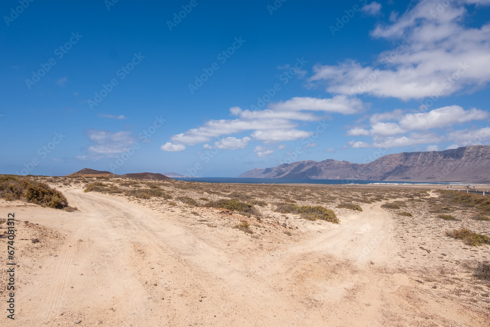 Desert landscape of white sand and desert bushes. Ocean and Famara cliff in the background. Dirt track. Sky with big white clouds. Lanzarote, Canary Islands, Spain.