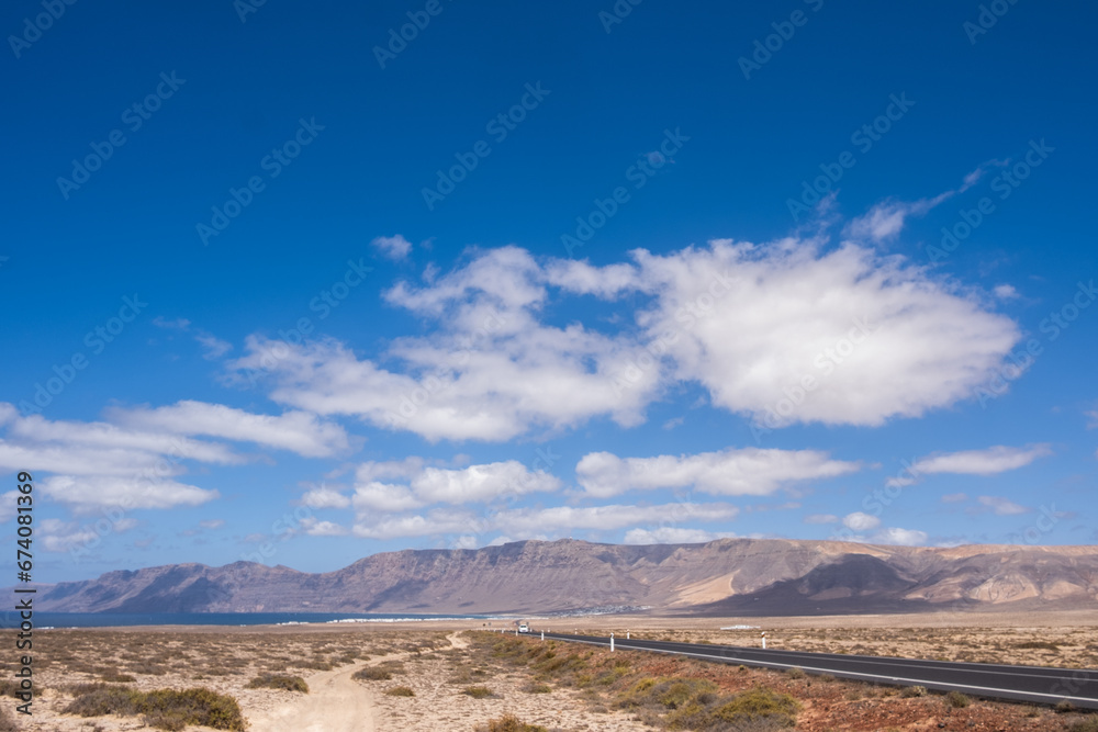 Obraz premium Desert landscape of white sand and desert bushes. Ocean and Famara cliff in the background. Dirt track. Sky with big white clouds. Lanzarote, Canary Islands, Spain.
