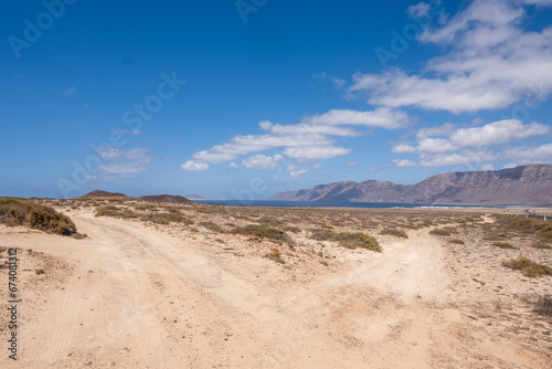 Desert landscape of white sand and desert bushes. Ocean and Famara cliff in the background. Dirt track. Sky with big white clouds. Lanzarote, Canary Islands, Spain.