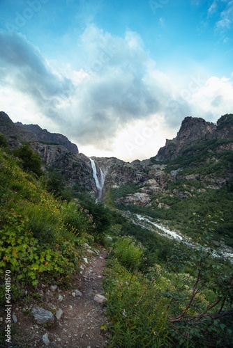 An incredible view of a majestic Shdugra waterfall in the distance, high in the Caucasus Mountains, rocky slopes, a rushing stream. Green herbs. Mazeri, Svaneti, Georgia. Vertical photo