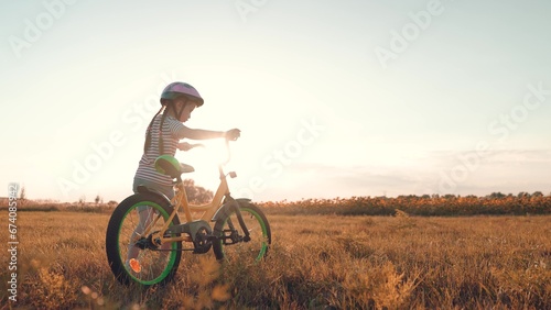 Little girl in helmet with small bicycle walks across dusk rural field
