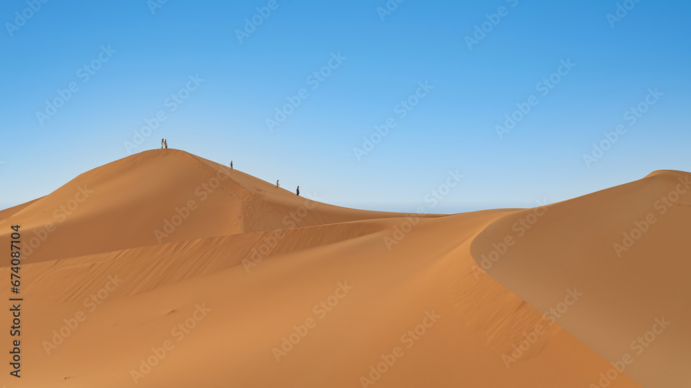 Unidentified people walking on sand dunes of Sahara Desert in northern Africa, a vast expanse of sand and dunes that stretches as far as the eye can see.