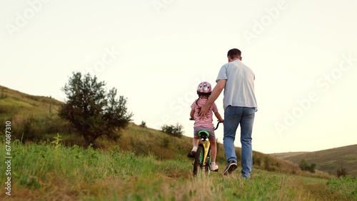 Attentive father teaches preschooler girl to ride bicycle in sunset field