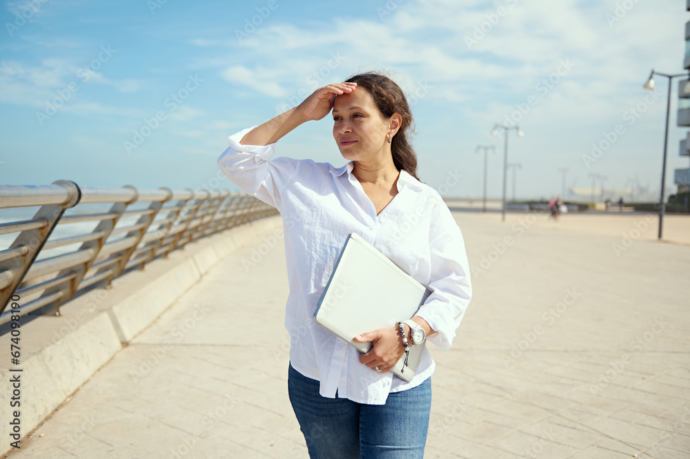 Confident young woman freelancer entrepreneur holding laptop and looking into the distance, standing on the promenade Confident young woman freelancer entrepreneur holding laptop and looking into the distance, standing on the promenade