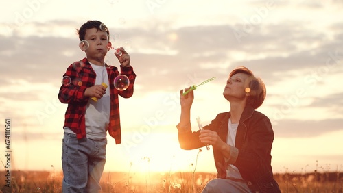 Mother with son blows soap bubbles in field under bright sunset low angle shot