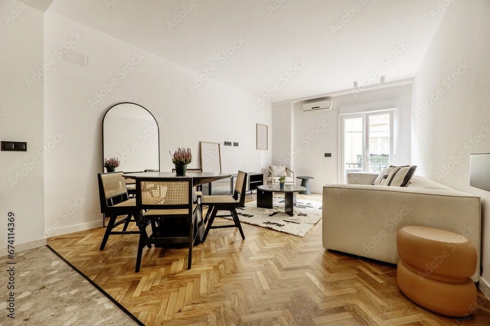 Fototapeta premium Living room of a modern house with circular black wooden table, chairs with matching backs and seats, mirrors leaning on the wall, a long sofa upholstered in beige fabric