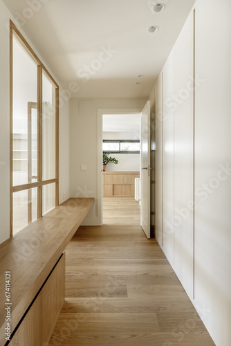 Corridor of a renovated apartment with smooth walls painted white, skylights and glass walls, a white work aramiro and an elongated oak plywood bench