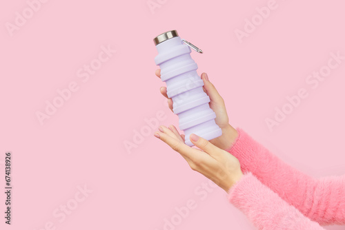 Womans hands holding collapsible reusable lilac water bottle on pink background. Sustainable lifestyle photo