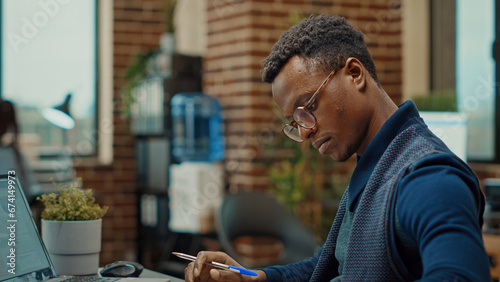 Corporate assistant taking notes on files, reviewing business documents and statistics to create new research plan. Young man in coworking space examining annual reports with information.
