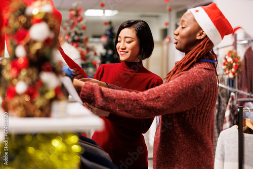 Store clerk helping asian client to find right measurement, recommending clothes and accessories for christmas dinner preparations at mall. Diverse women discussing about fabrics quality. photo