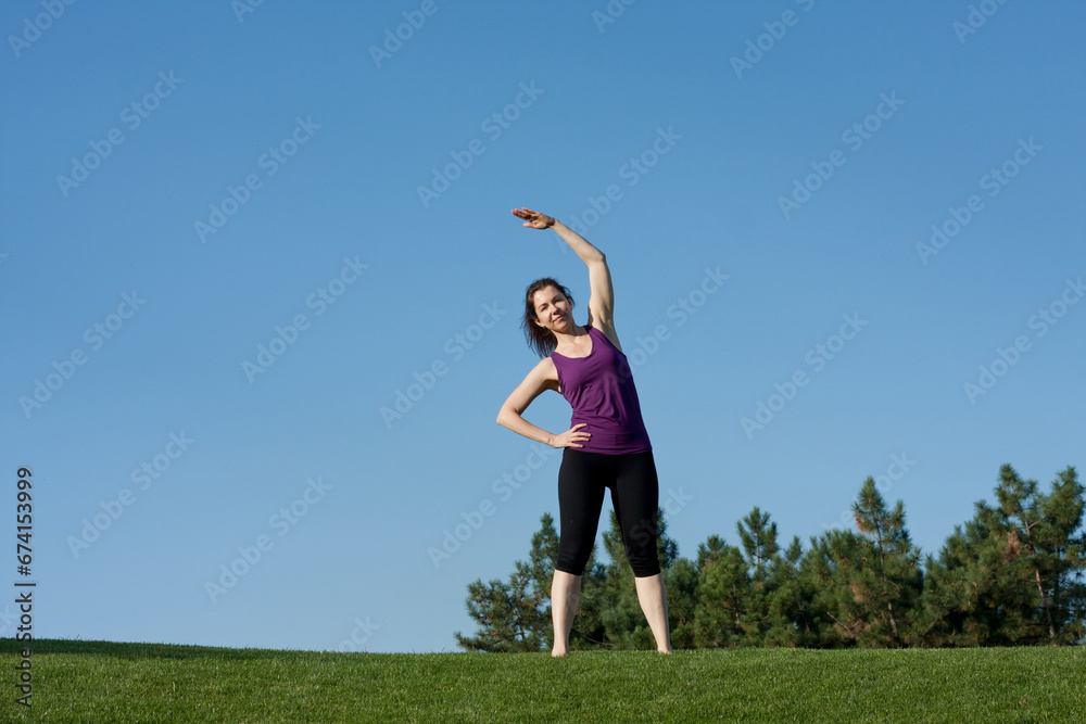 Athletic young woman doing warm-up before fitness workout in park, blue sky background. beautiful woman practices yoga, Pilates, standing barefoot on grass, bending body to side. Copy space Athletic young woman doing warm-up before fitness workout in park, blue sky background. beautiful woman practices yoga, Pilates, standing barefoot on grass, bending body to side. Copy space