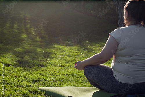 Portrait of overweight Caucasian middle-aged woman practicing yoga in nature. Fat woman meditating, relaxing in lotus position, sitting on fitness mat. Calm, relaxed, zen, stop stress. View from back