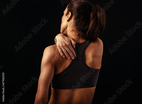 Young strong sporty woman suffering from pain in shoulder in sport wear. Touching and massaging the hand. Sports exercising injury. Closeup portrait dark black background