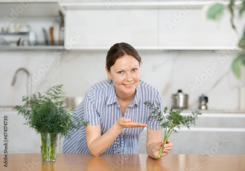 Youthful housewife stands in kitchen  clutching vase filled with vibrant bunch of dill weed and fennel. She took out of bouquet and shows fluffy sprig of dill