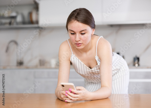 Pretty young girl leaning on kitchen table and looking at screen of her smartphone with interest