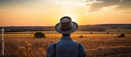 A person wearing a hat glasses and holding a camera is positioned in a field during the sunset
