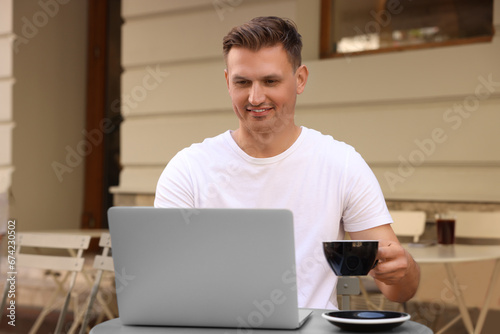 Handsome man with cup of drink working on laptop at table in outdoor cafe