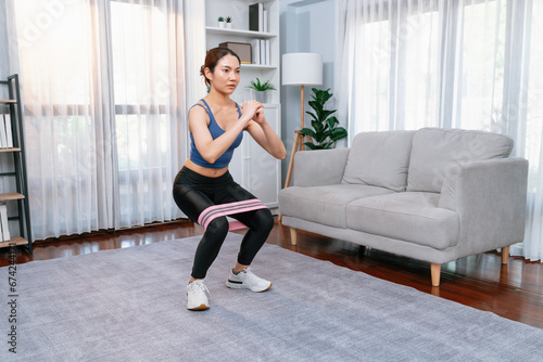 Vigorous energetic woman doing exercise at home with resistance sport band for leg muscle gain. Young athletic asian woman strength and endurance training session as home workout routine with squat.