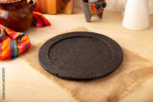 Volcanic stone comal, traditional kitchen utensil used in Mexican gastronomy, in the background stone molcajete and clay pot with typical Mexican fabric on wooden table.