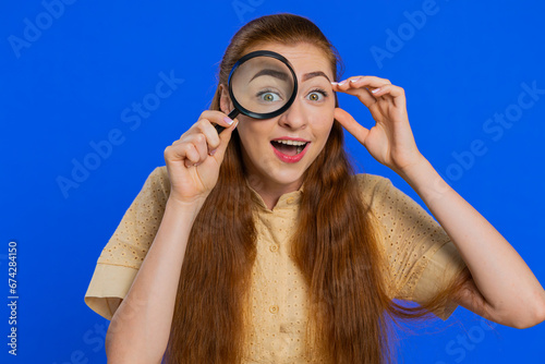Investigator researcher scientist woman holding magnifying glass near face  looking into camera with big zoomed funny eye  searching  analysing. Pretty girl isolated on blue studio background indoors